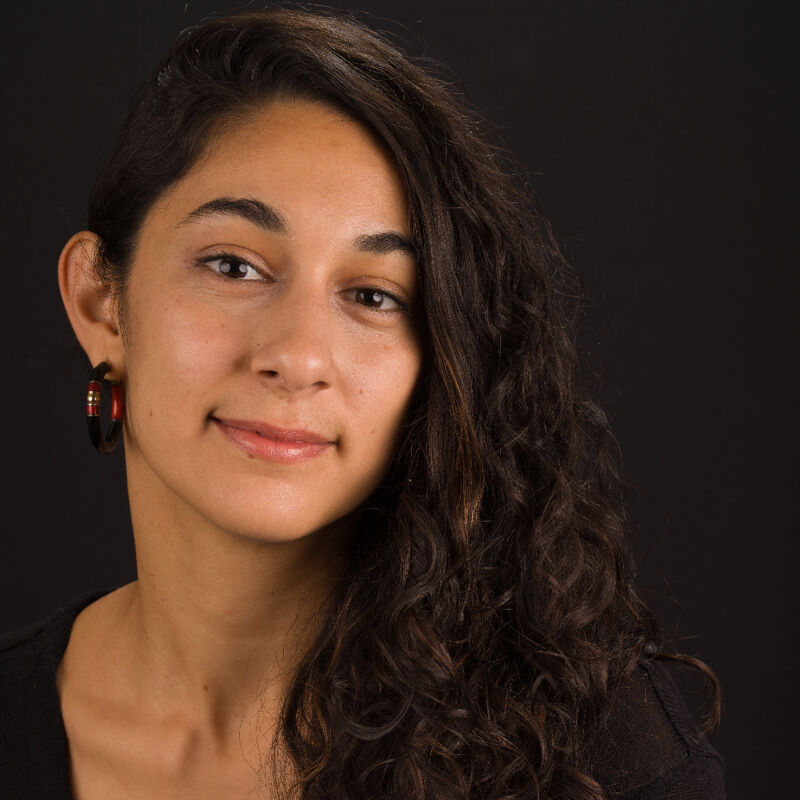 The image shows a woman with long, dark, curly hair and brown eyes. She is wearing a black top and hoop earrings. The background is a solid black. She is looking at the camera with a slight smile.